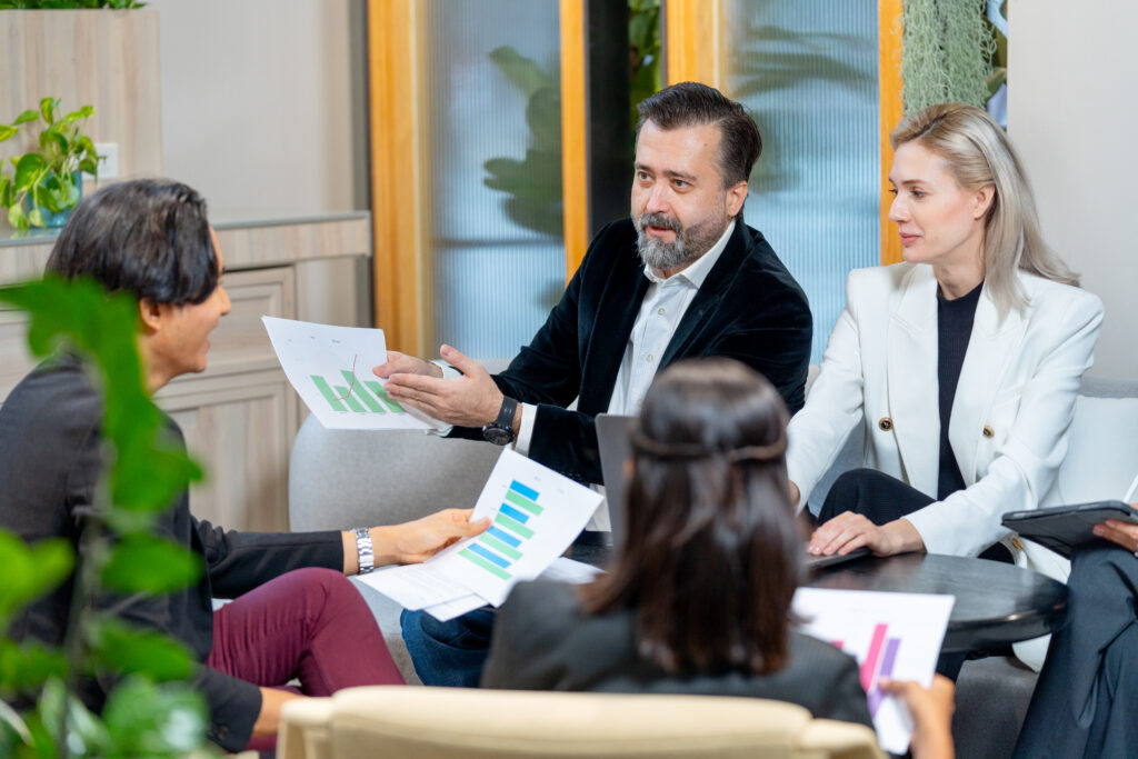 A group of four people sitting together in a modern office setting, engaging in a discussion about pricing accounting services. One man in a velvet blazer is gesturing, while others listen attentively. Plants and office furniture are visible in the background.