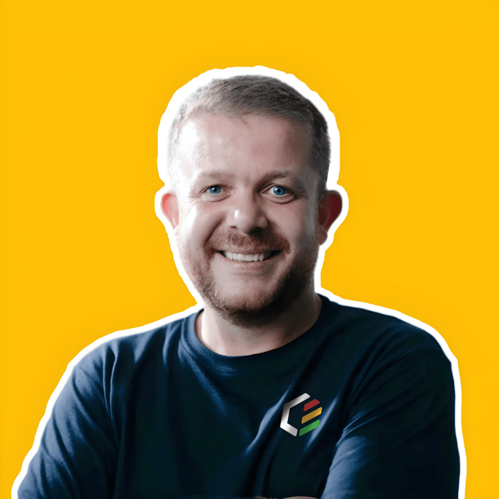 A person with short hair and a beard is smiling at the camera. They are wearing a dark shirt with a small multicolored logo on the chest. The background is a bright yellow, and there is a white outline around the person.