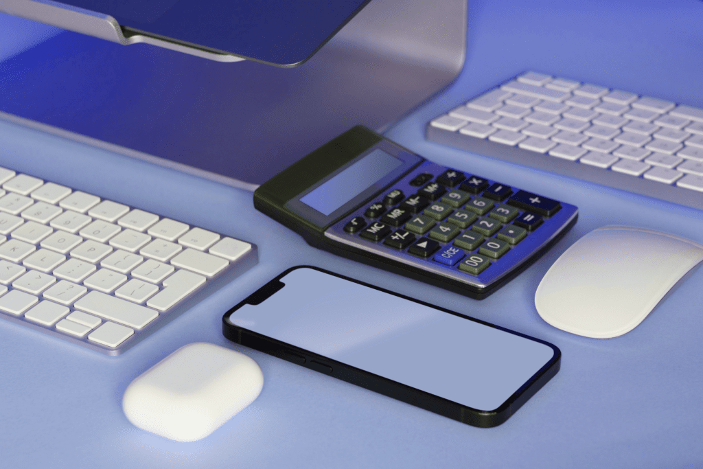 A desk setup with a computer monitor, two keyboards, a mouse, a calculator for resilient accounting, a smartphone, and wireless earbuds, all arranged neatly on a blue surface.