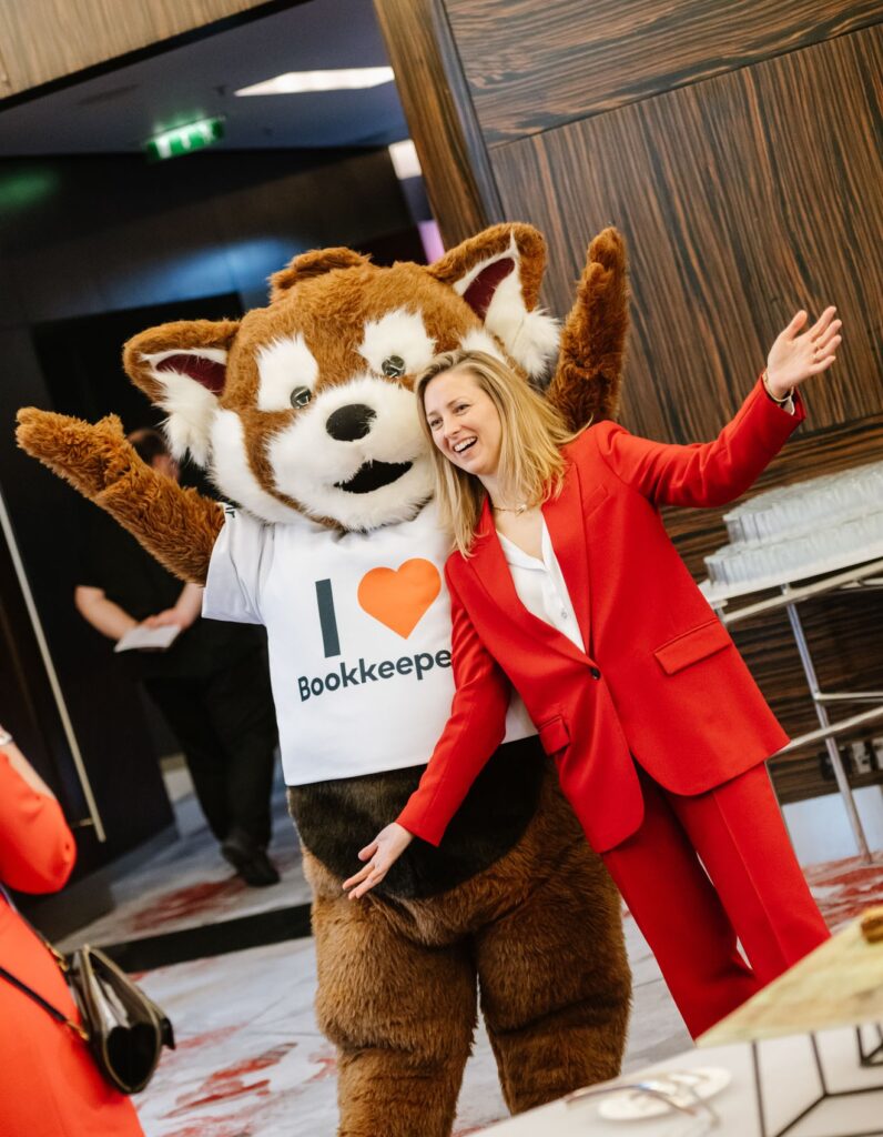 Ami Copeland, dressed in a vibrant red suit, poses happily with a costumed mascot wearing a shirt that says I ♥ Bookkeepers. They stand inside a modern building adorned with wood-paneled walls and a light floor.