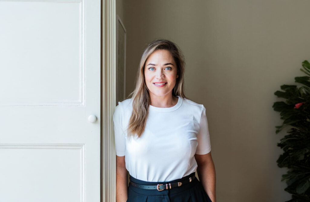 A person with long hair is leaning against a white wall, wearing a white shirt and dark pants with a belt. They are smiling and the room has soft lighting. A plant is partially visible in the background.