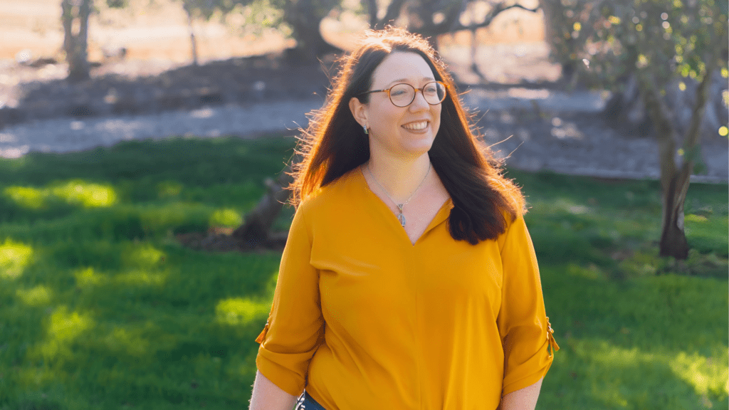 Karen Reyburn, a woman with long dark hair and glasses, stands smiling in a sunlit outdoor setting. Her yellow blouse contrasts beautifully with the lush green grass and trees in the background.