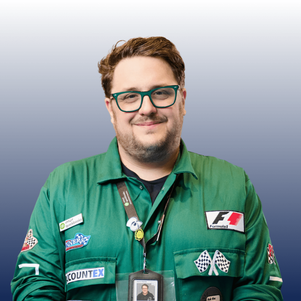 A man, identified by his ID badge as Aaron Patrick, wearing glasses and a green Formula 1 jacket adorned with various patches, stands against a gradient background. He is smiling confidently at the camera.