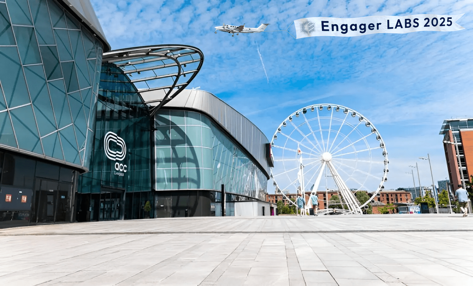 Modern building with glass facade labeled OCC next to a large Ferris wheel under a blue sky. A plane flies overhead trailing a banner that reads Engage LABS 2025.