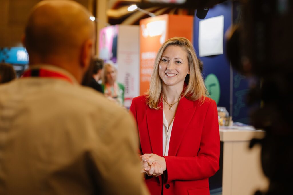 Ami Copeland, dressed in a bright red blazer, smiles warmly during a conversation with someone whose back is to the camera. They are in a well-lit indoor setting adorned with colorful signs and displays.