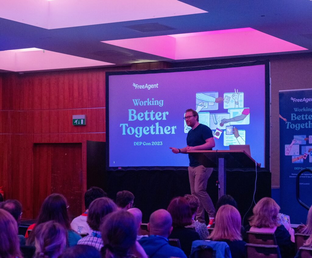 Stu McEwan on stage in front of an audience at DEP Con 2023. A large screen displays Working Better Together under the FreeAgent logo. The room is filled with seated attendees, and the lighting is a mix of blue and purple tones.