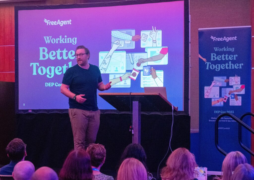 Stu McEwan presents at the FreeAgent Working Better Together event. They stand on stage next to a lectern, with a slide displayed on a large screen behind them, and several attendees visible in the audience.