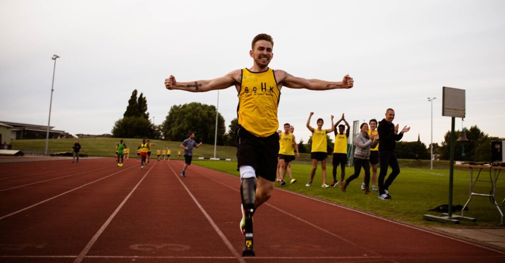 Andy Grant, with a prosthetic leg triumphantly crosses the finish line on a red track, arms outstretched. Spectators and fellow runners in yellow shirts cheer him on from the sidelines.