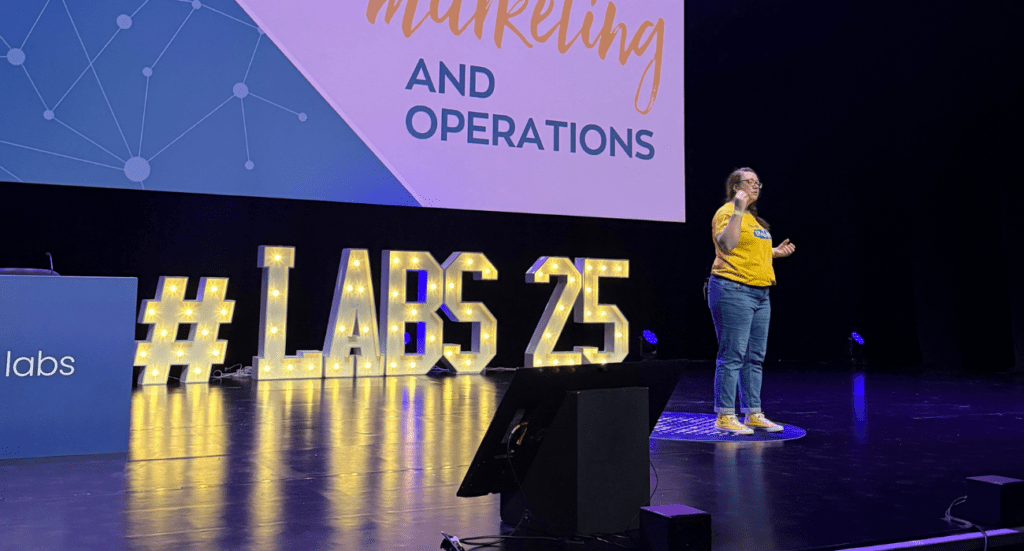 Karen Reyburn of We Are PF stands on stage giving a presentation. Clad in a yellow shirt and jeans, they stand before large illuminated letters spelling #LABS 25. The screen displays Marketing and Operations, emphasizing how these teams work together seamlessly.