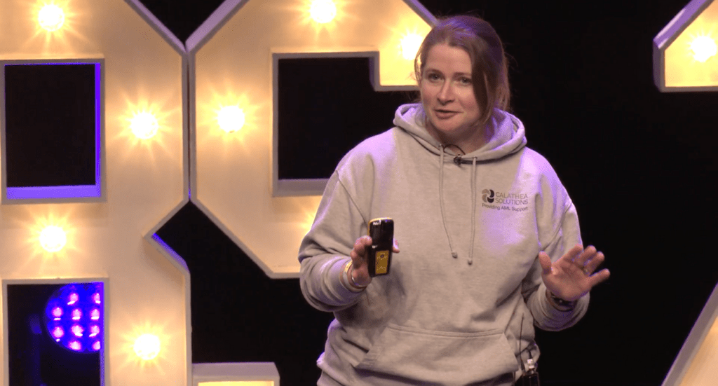 Lucy Brown in a gray hoodie speaks on stage, holding a small device. Behind her, large illuminated letters and numbers spell out Compliance. The setting appears to be a conference focused on the latest trends in technology and regulation.