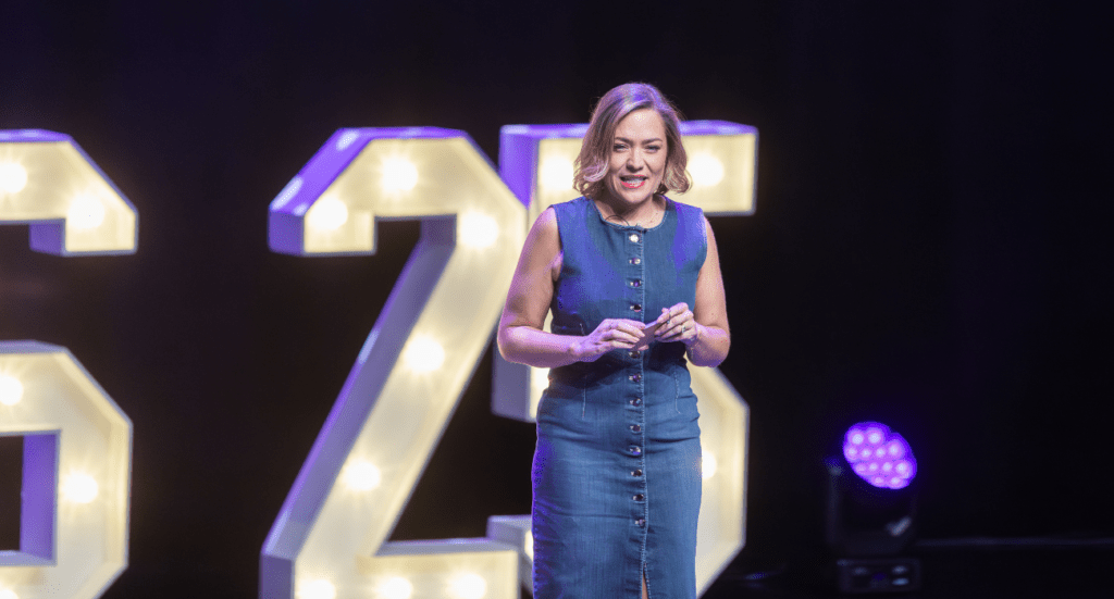 Jo Wood of The 6 Figure Bookkeeper stands on stage holding a microphone, wearing a sleeveless denim dress. Behind her is a large, illuminated number 25, celebrating Jo Woods legacy as a trusted business leader. The stage lights enhance the anniversary events sense of achievement and joy.