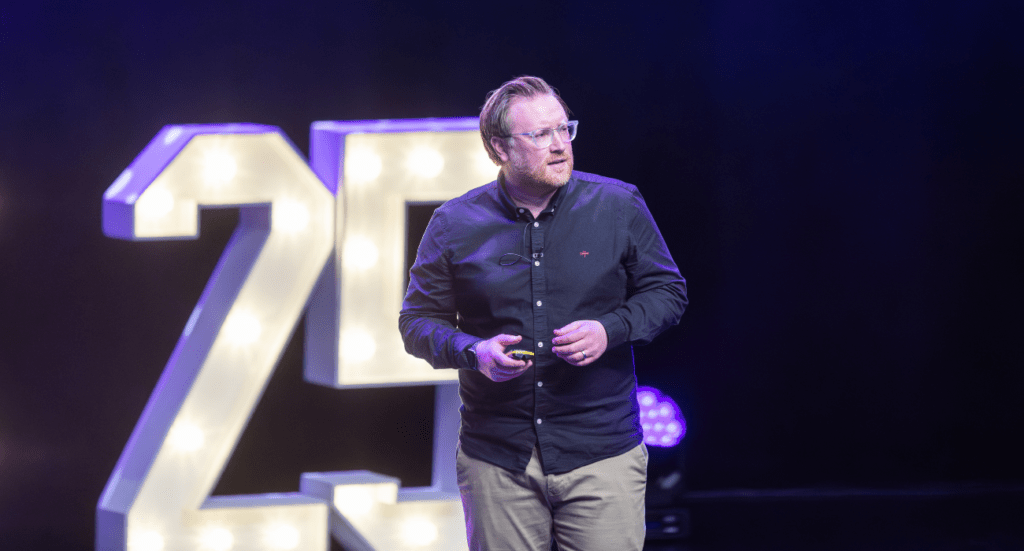 Stu McEwan of FreeAgent, with glasses and a beard, dons a dark shirt and khaki pants as he addresses the audience on stage. A large illuminated 25 shines behind him, symbolizing the elephant in the room—the opportunity in change.