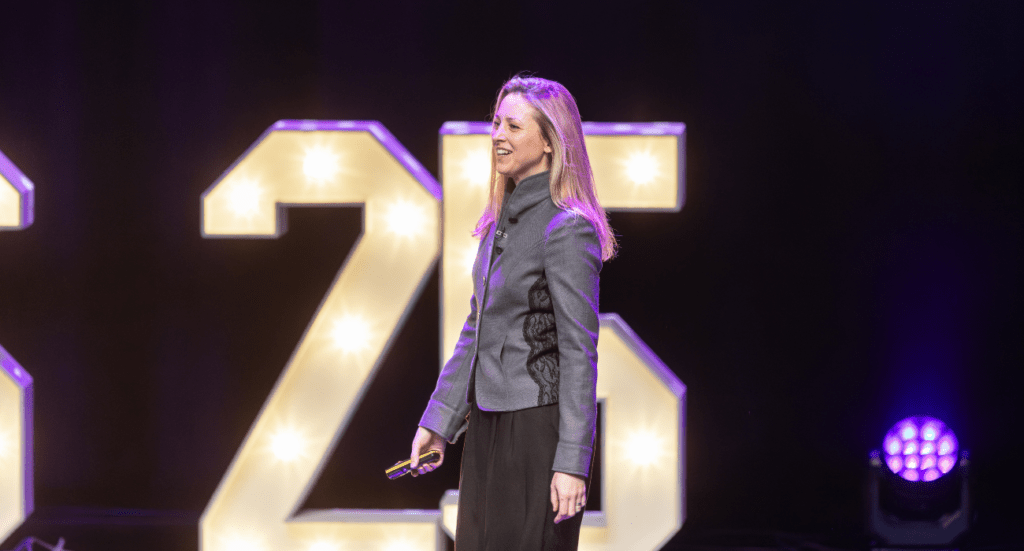 Ami Copeland, CEO of the Institute of Certified Bookkeepers (ICB) is speaking on stage in front of large, illuminated numbers 25, discussing the future of Tax Digital. The dark background and purple stage lights create a captivating glow. The individual, holding a microphone, is dressed in a gray jacket and black pants.